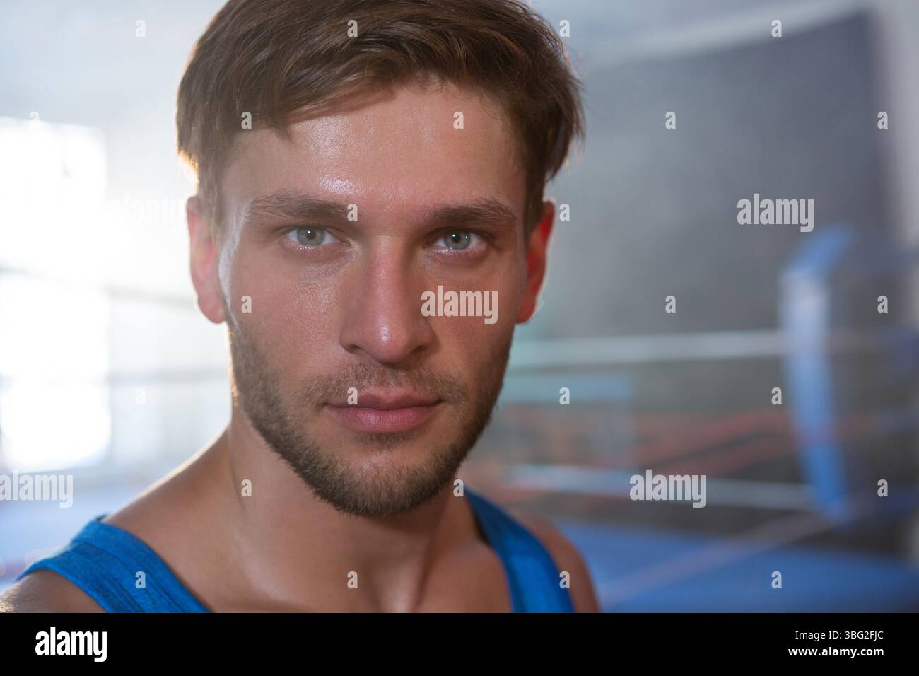 Atleta in piedi sul ring di boxe con canotta blu che guarda la fotocamera con una borsa da pugno Foto Stock