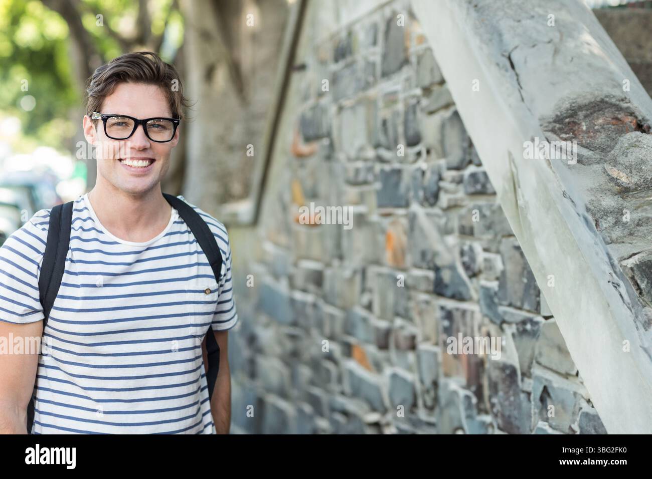 Uomo in piedi accanto a un muro di pietra sul marciapiede con zaino con occhiali da vista a righe, spazio copia Foto Stock