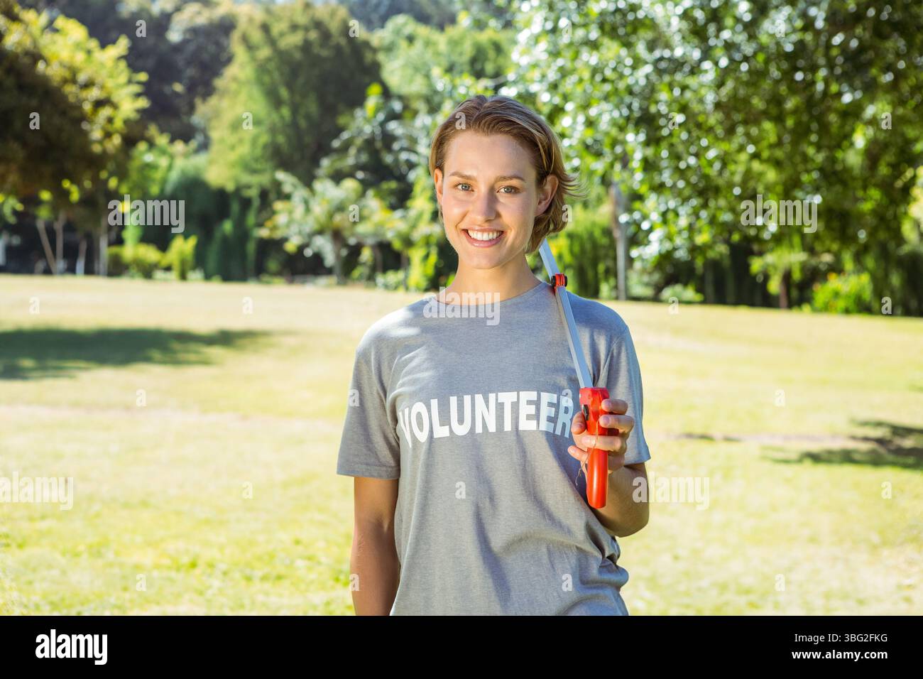 Donna sorridente in piedi all'aperto che indossa una t-shirt VOLONTARIA e tiene in mano uno strumento per la raccolta di rifiuti rossi Foto Stock