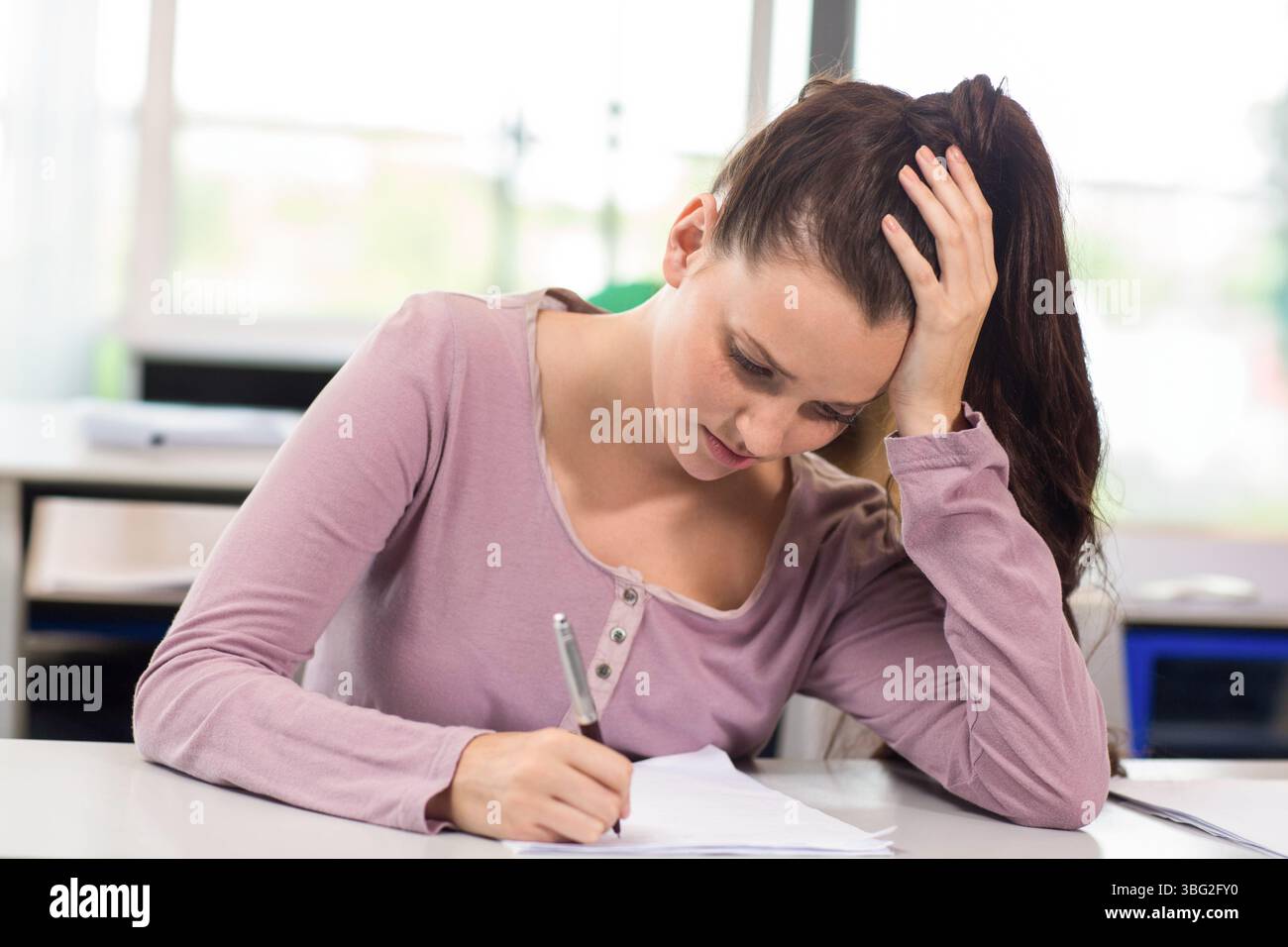 Donna che indossa una scritta a manica lunga in malva con penna su carta alla scrivania dell'aula mentre riposa la testa Foto Stock