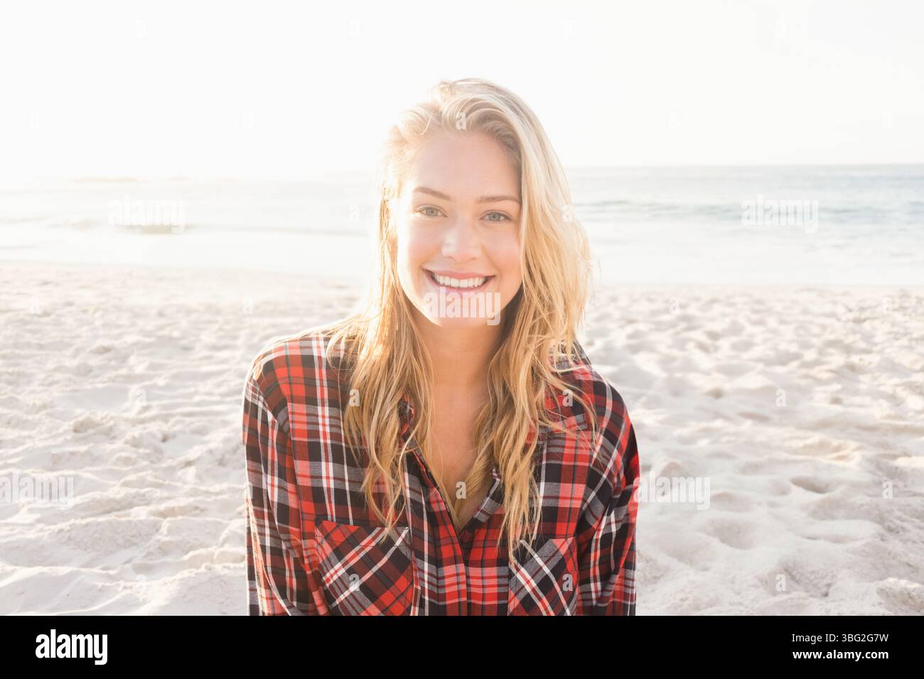 Donna sorridente in piedi su una spiaggia sabbiosa vicino al bordo dell'acqua e indossa una camicia a quadri sotto la luce del sole retroilluminato Foto Stock