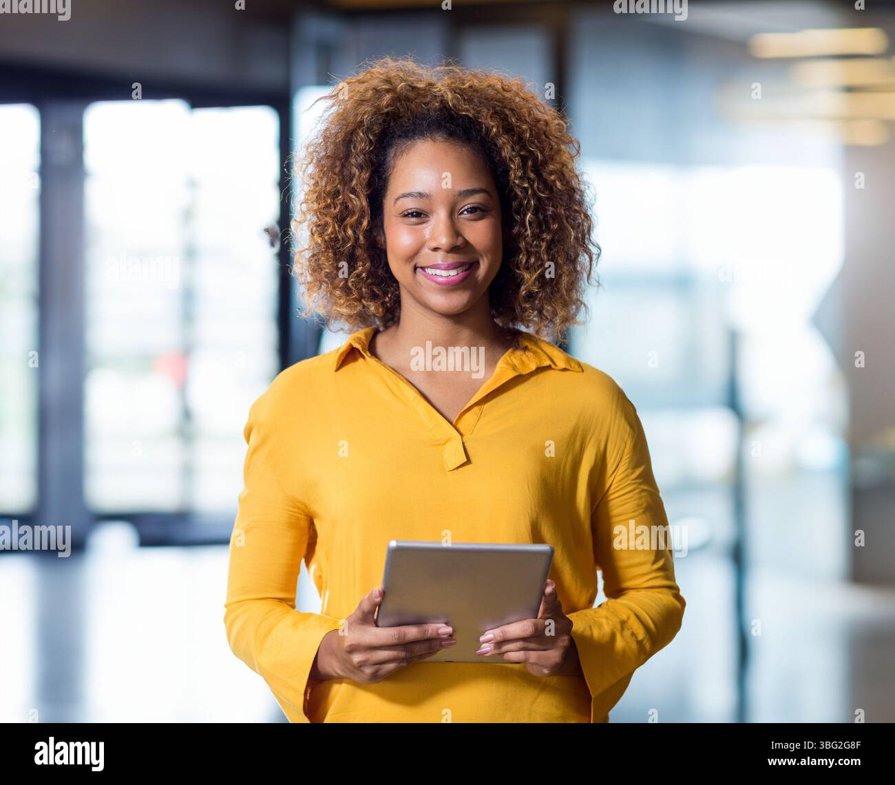 Donna afroamericana in piedi nell'atrio dell'ufficio con camicetta gialla senape e tablet argento Foto Stock