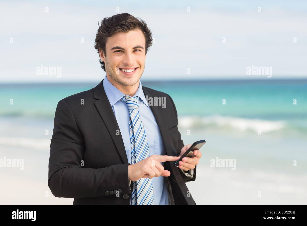 Uomo in viaggio di lavoro in piedi su una spiaggia sabbiosa che balla le dolci onde turchesi e tiene in mano lo smartphone Foto Stock