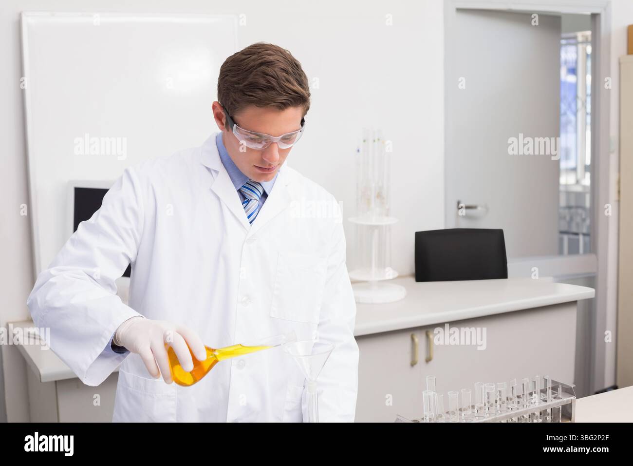 Matraccio di Erlenmeyer che versa una soluzione di colore giallo brillante attraverso l'imbuto in vetro nel becher sul banco da laboratorio bianco Foto Stock