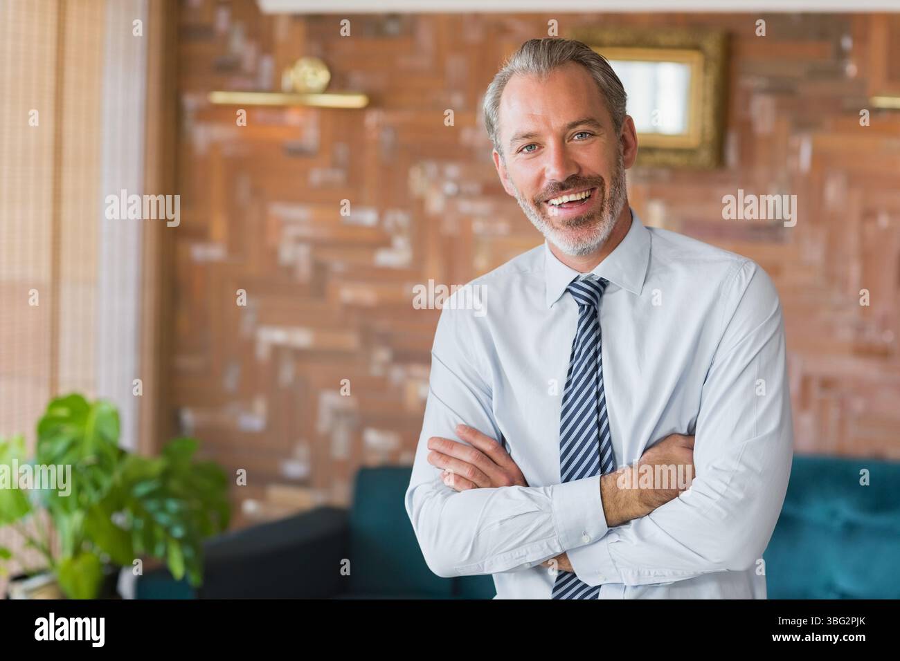 Uomo d'affari in camicia e cravatta con le braccia piegate nel salotto con pannelli in legno e divano color tè Foto Stock