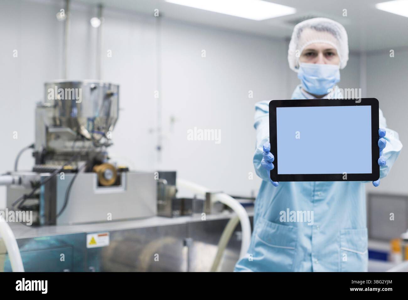 Scienziato caucasico uomo in tuta da laboratorio blu e guanti mostrano compressa vuota, macchine sullo sfondo Foto Stock