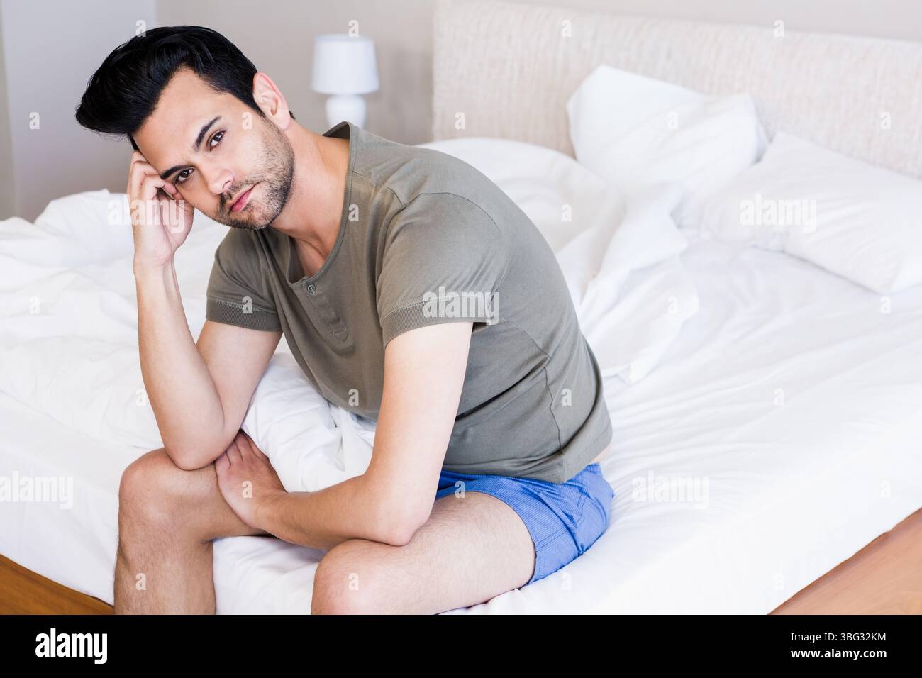 Uomo medio adulto seduto sul bordo di un letto non fatto in camera da letto con lampada per comodino che tiene la testa Foto Stock