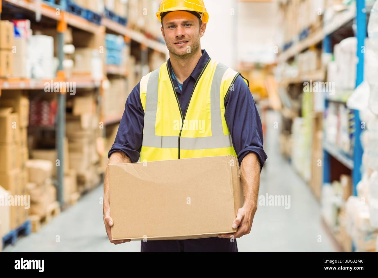 Lavoratore di magazzino che indossa elmetto e giubbotto che tiene la scatola di cartone nella corsia tra gli scaffali in metallo Foto Stock