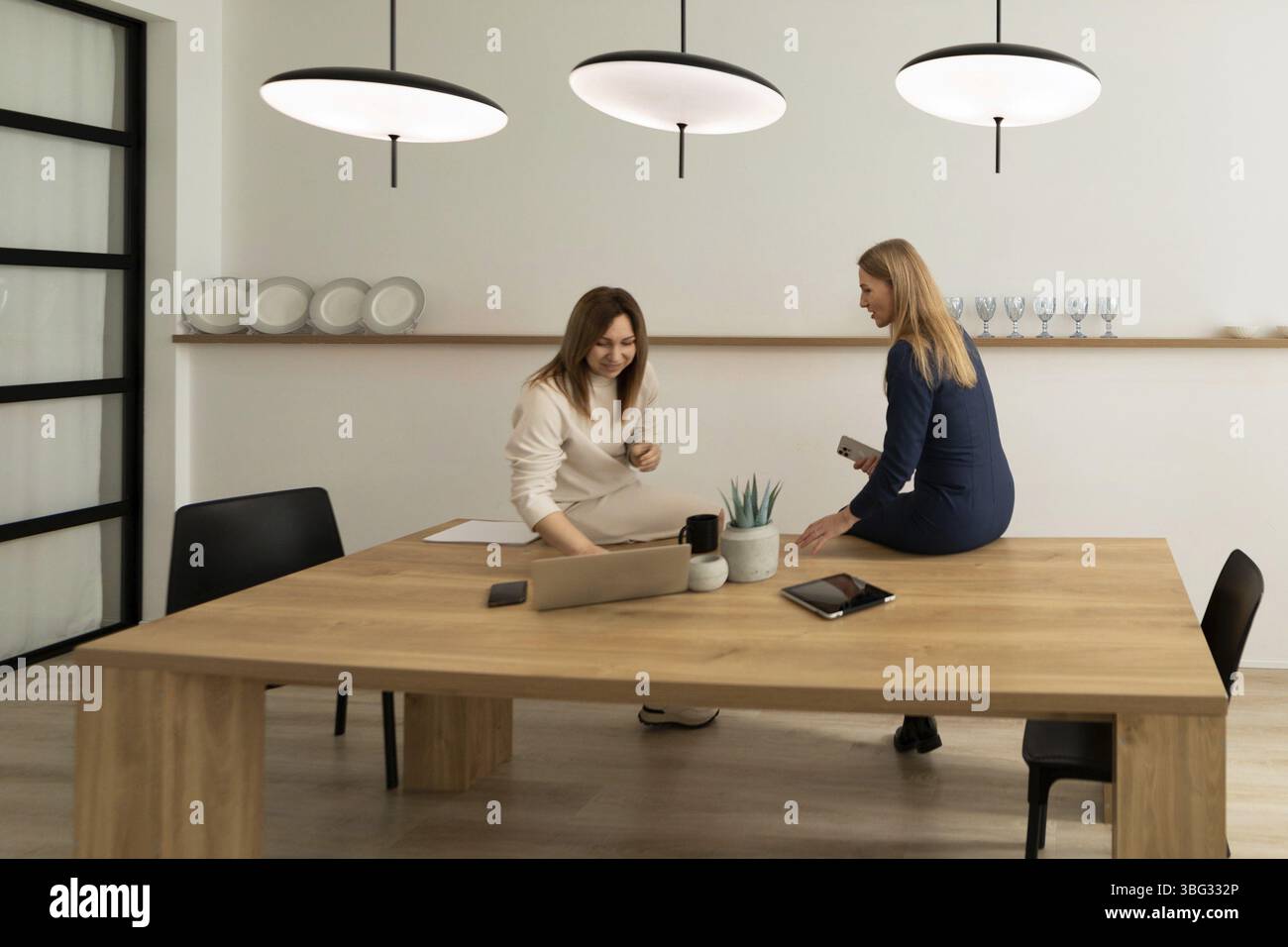 Due donne stanno scambiando idee su un tavolo di legno contemporaneo in un elegante spazio di lavoro. Uno è seduto sul tavolo mentre l'altro è appoggiato contro di esso, IND Foto Stock