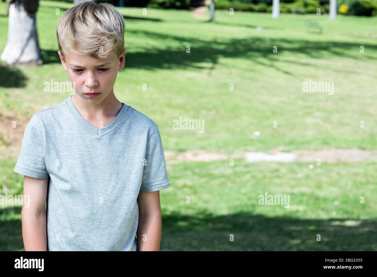 Ragazzo in piedi sull'erba tagliata nel parco, guardando verso il basso con espressione triste, copia spazio Foto Stock