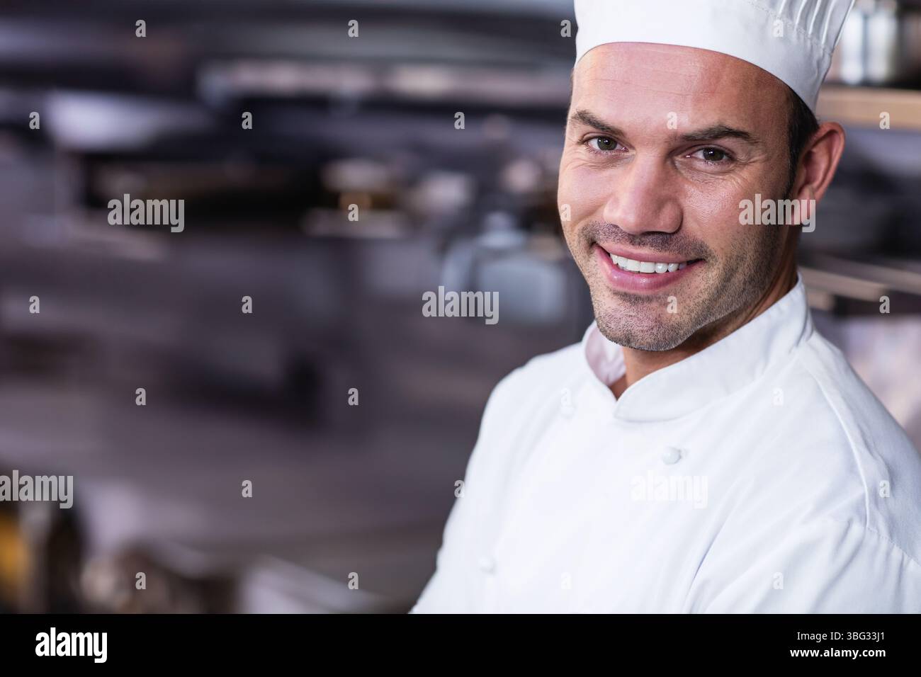 Lo chef maschile sorride verso lo spettatore nella cucina del ristorante con giacca e cappello bianchi dello chef, spazio copia Foto Stock