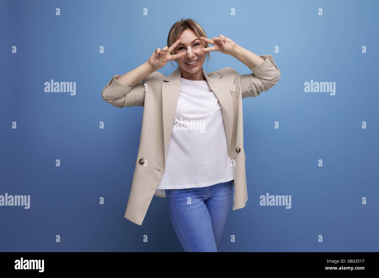 Nahaufnahme einer positiven blondn jungen frau in einer beige jacke mit emoionen auf ihrem gesicht im studio Foto Stock