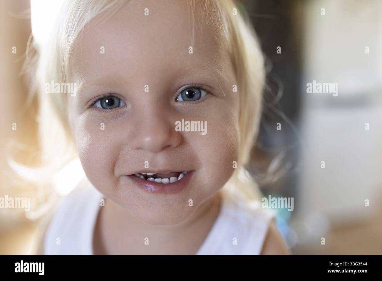 Un bambino allegro con i capelli biondi e gli occhi azzurri sorride molto mentre gioca in casa. La calda luce del sole illumina delicatamente il canale Foto Stock
