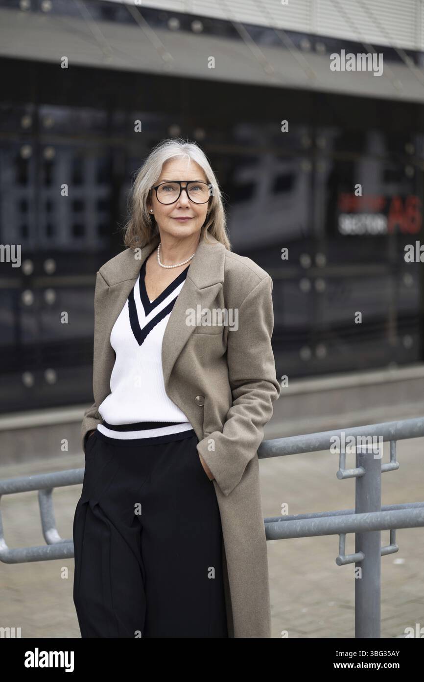 Una donna anziana alla moda è orgogliosa di stare accanto a una ringhiera, vestita con un elegante outfit con un maglione con scollo a V e un cappotto lungo, Rade Foto Stock