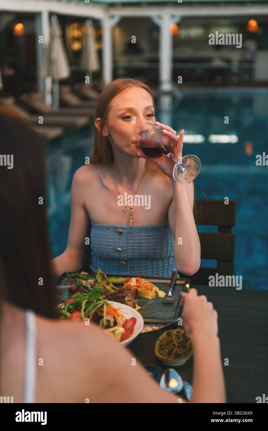 Una donna bionda, ventenne, gusta la cena con vino rosso in un ristorante dell'hotel, con la piscina e le luci soffuse della sera sullo sfondo. Foto Stock
