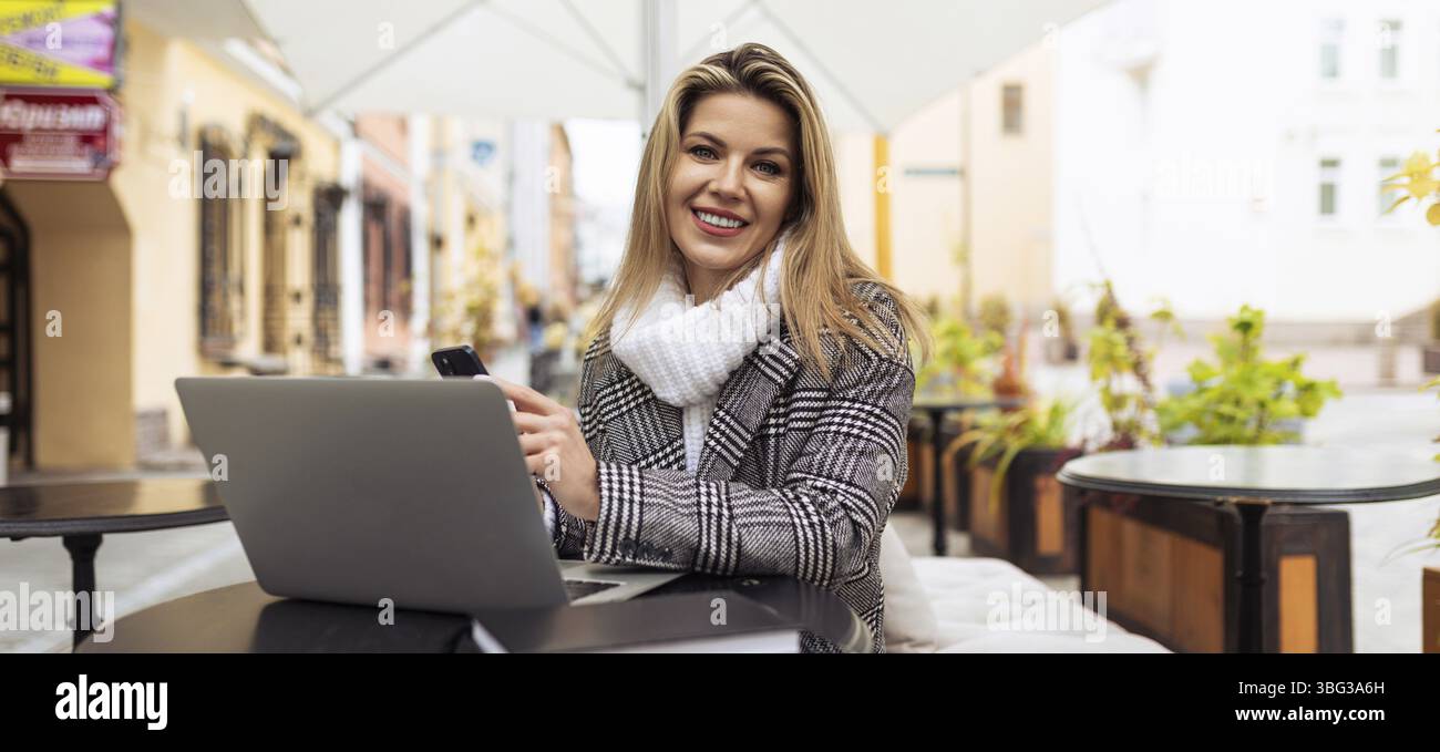 Junge bionda journalistin mit laptop sitzt auf der terrasse in einem Cafe Foto Stock