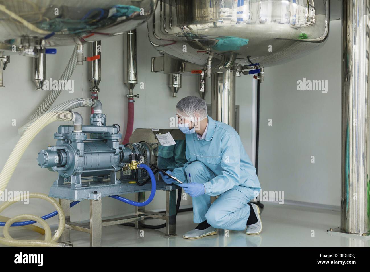 Scienziato in tuta da laboratorio e maschera antigas sotto grandi serbatoi in acciaio controllano il motore Foto Stock