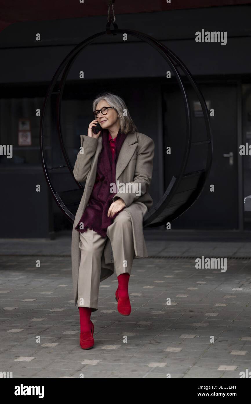 Una donna più anziana è seduta in un'altalena circolare e parla al telefono. Indossa un outfit elegante con calzini rossi, che si abbina a un dorso urbano contemporaneo Foto Stock