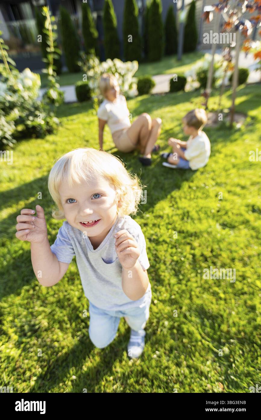 Una madre sta felicemente interagendo con i suoi due figli piccoli in un parco giochi. Un figlio corre avanti con un grande sorriso, mentre l'altro è seduto vicino Foto Stock