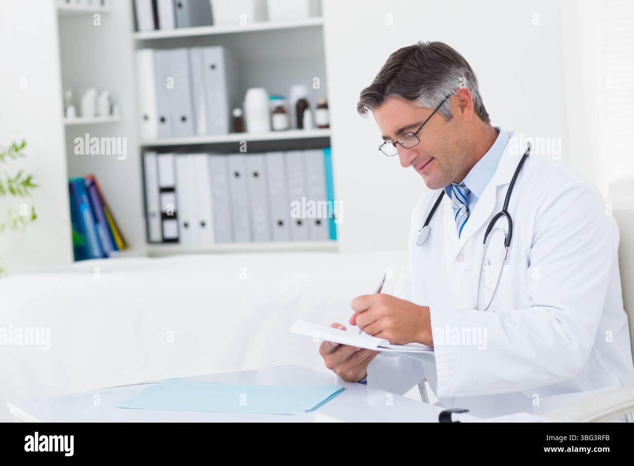 Maschio medico in bianco cappotto scrittura note su appunti a scrivania medica con stetoscopio, copia spazio Foto Stock