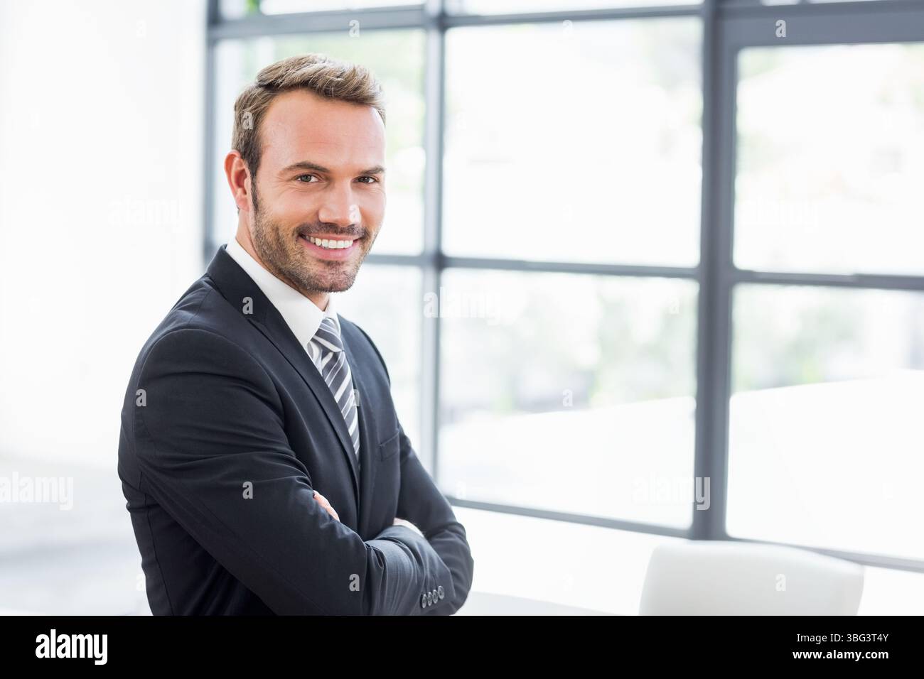 Uomo d'affari che indossa una tuta scura con bracci a righe e grandi finestre in un ufficio moderno Foto Stock