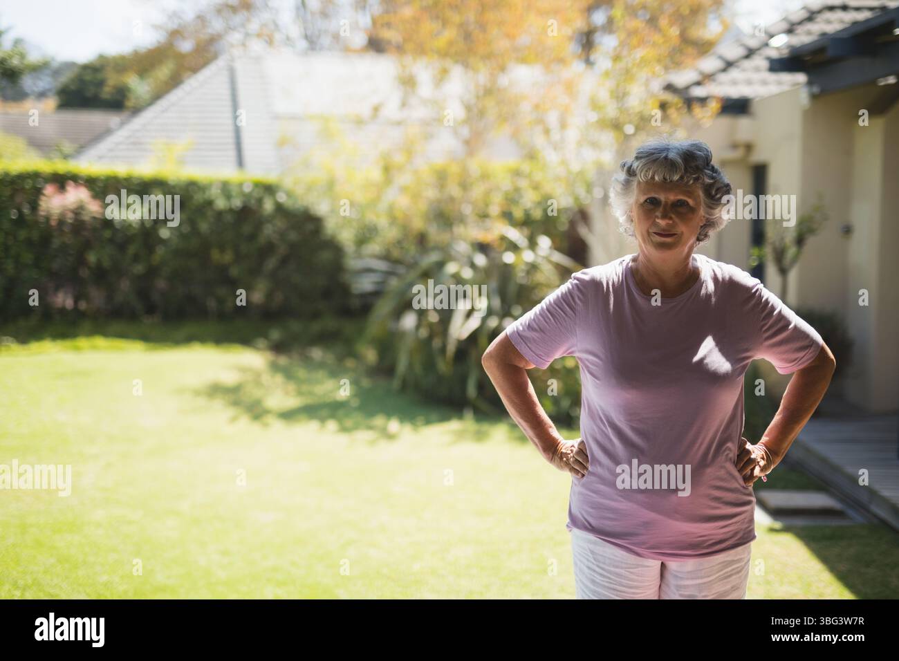 Donna anziana in piedi con le mani sui fianchi nel cortile di periferia, con siepe e ponte, copia spazio Foto Stock