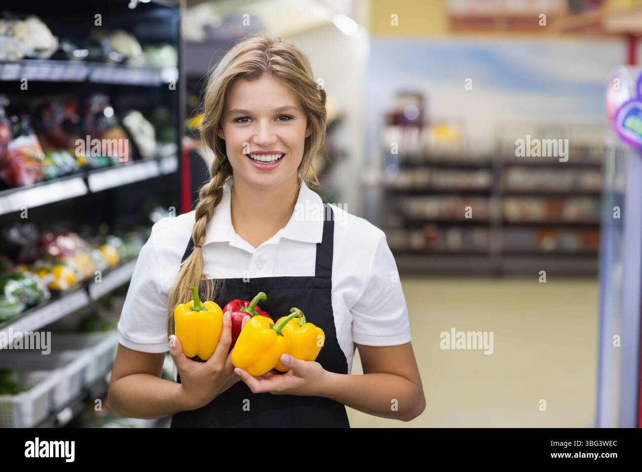 Lavoratrice sorridente che indossa un grembiule nero sulla polo nella corsia dei prodotti con peperoni Foto Stock