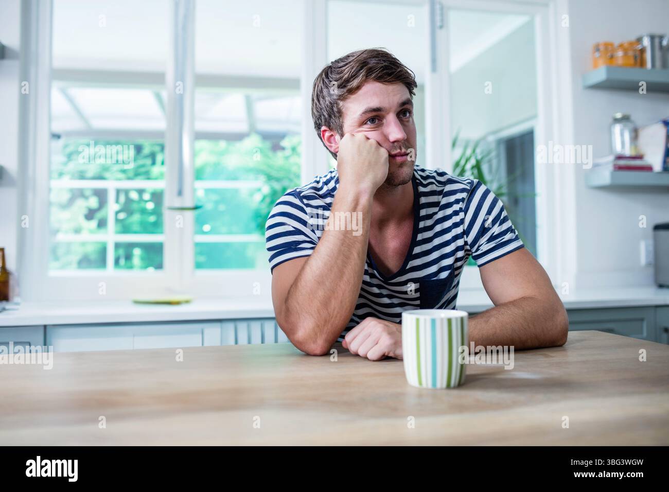 Uomo bianco appoggiato al gomito, che guarda verso destra al tavolo della cucina con tazza e grandi finestre Foto Stock