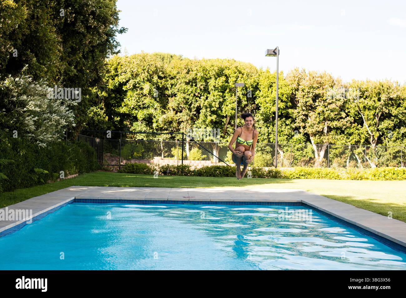 Donna di 20 anni che indossa un costume da bagno in due pezzi verde neon per tuck jump sopra il ponte della piscina in pietra del cortile posteriore Foto Stock