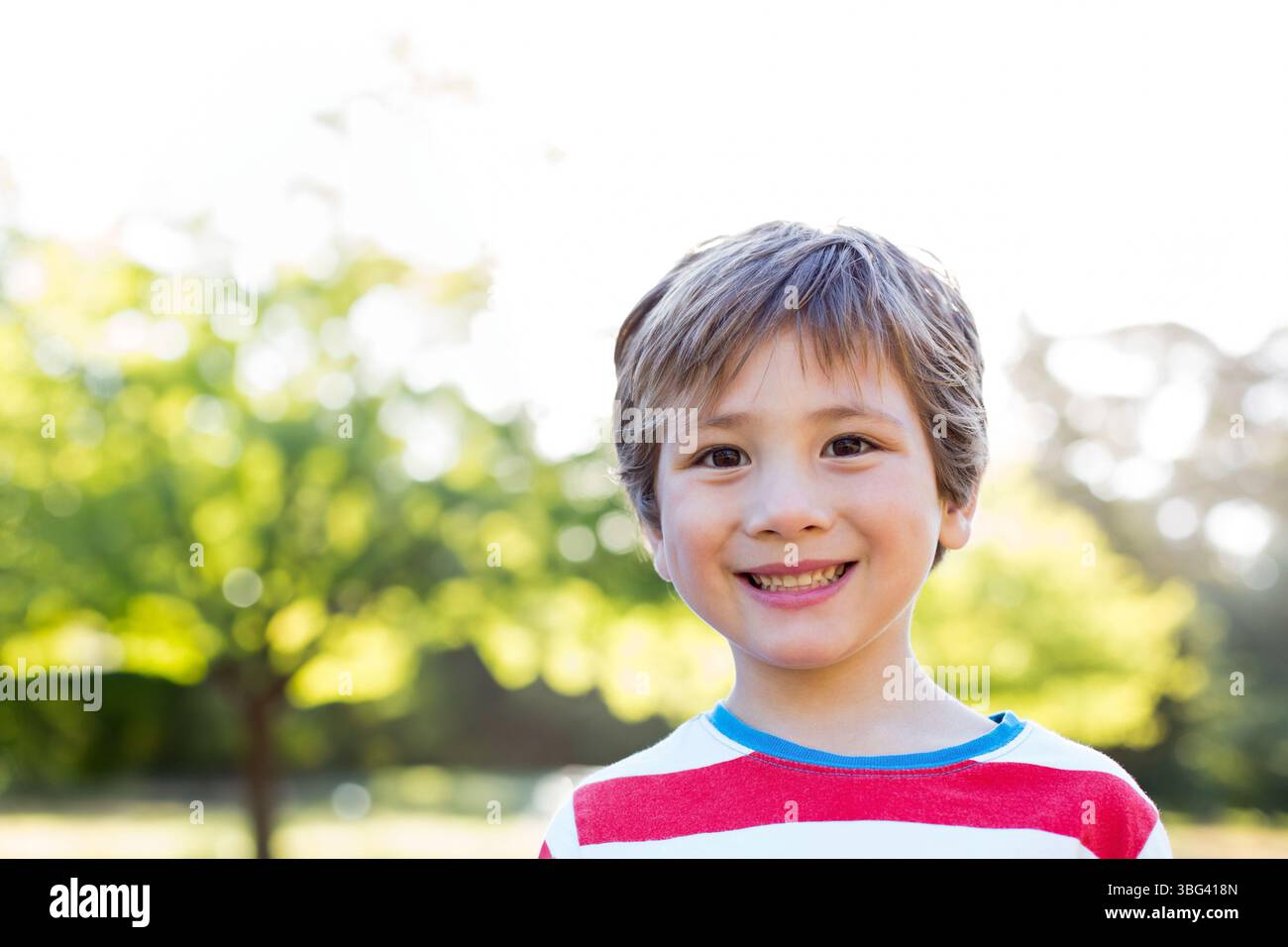 Bambino maschio in piedi e sorridente al parco con T-shirt a righe rosse e bianche sotto la luce del sole Foto Stock