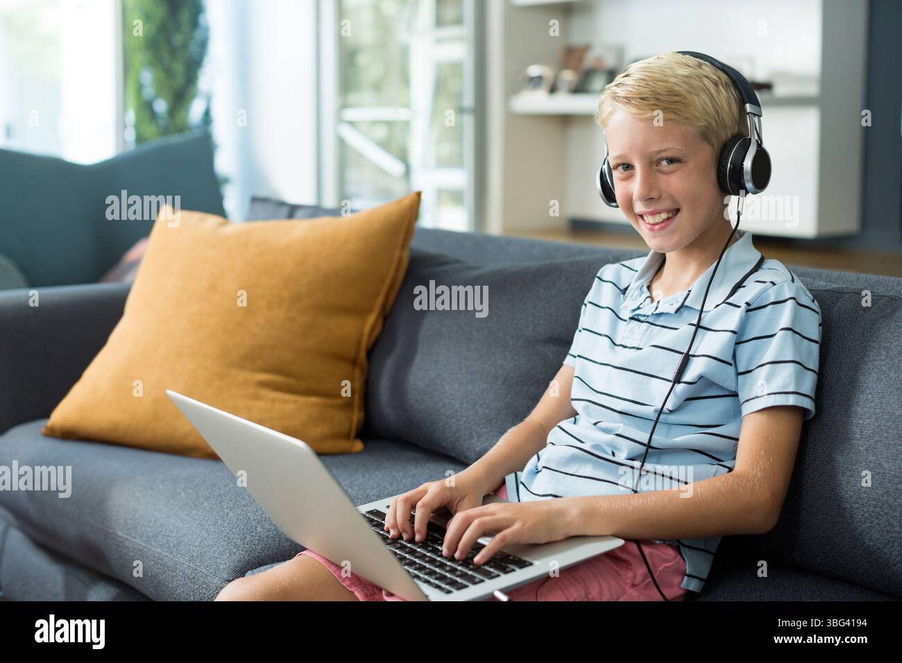 Ragazzo che scrive sul computer portatile indossando polo a righe e cuffie sul divano di casa con cuscini Foto Stock