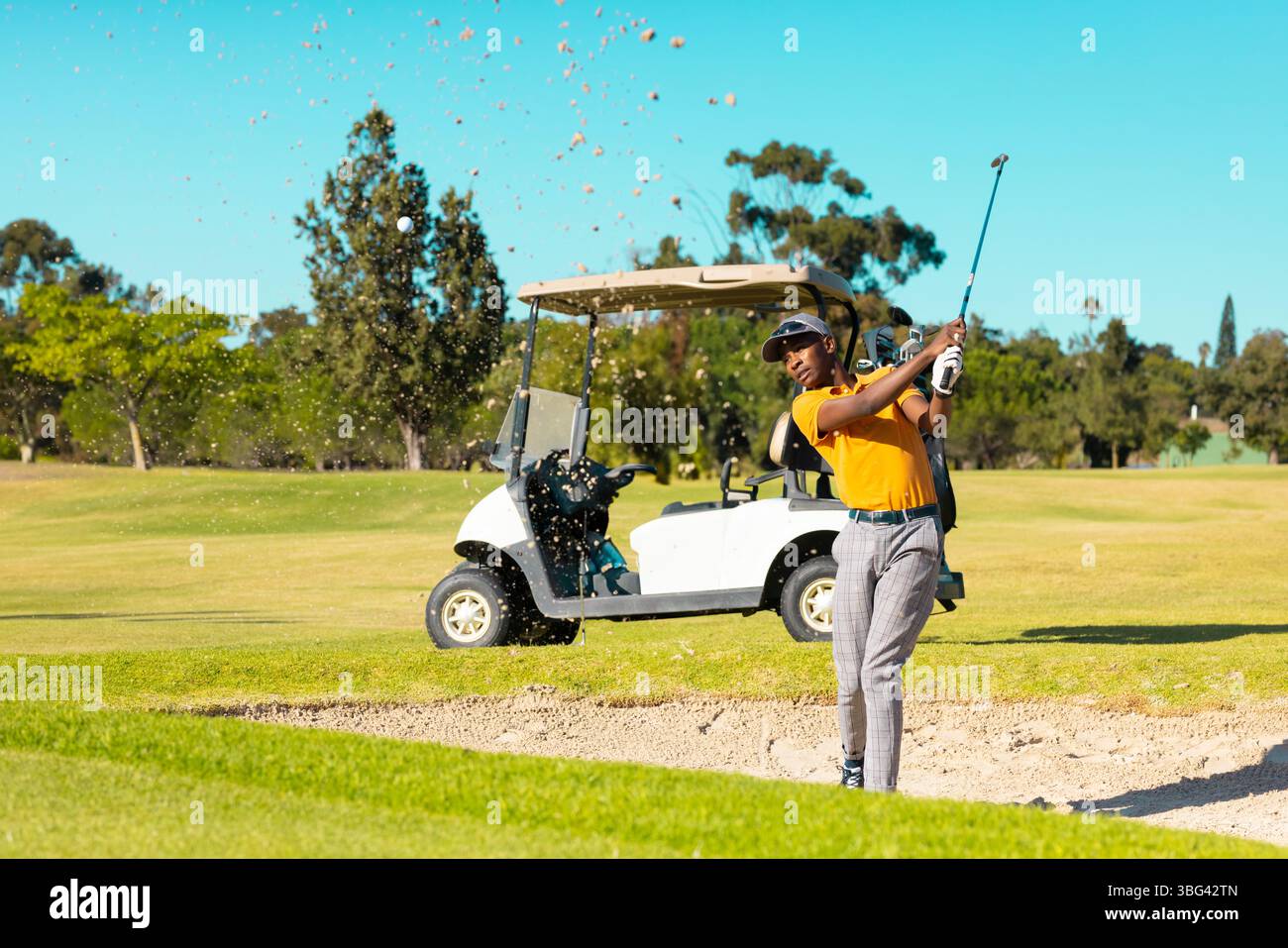 Golfista afro-americano che oscilla dal bunker di sabbia che invia palla in polo arancione, copia spazio Foto Stock