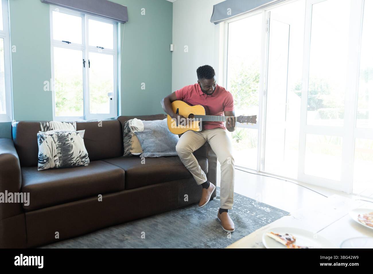 Uomo afroamericano che suona la chitarra acustica su un divano in pelle nel soggiorno accanto a fette di pizza Foto Stock