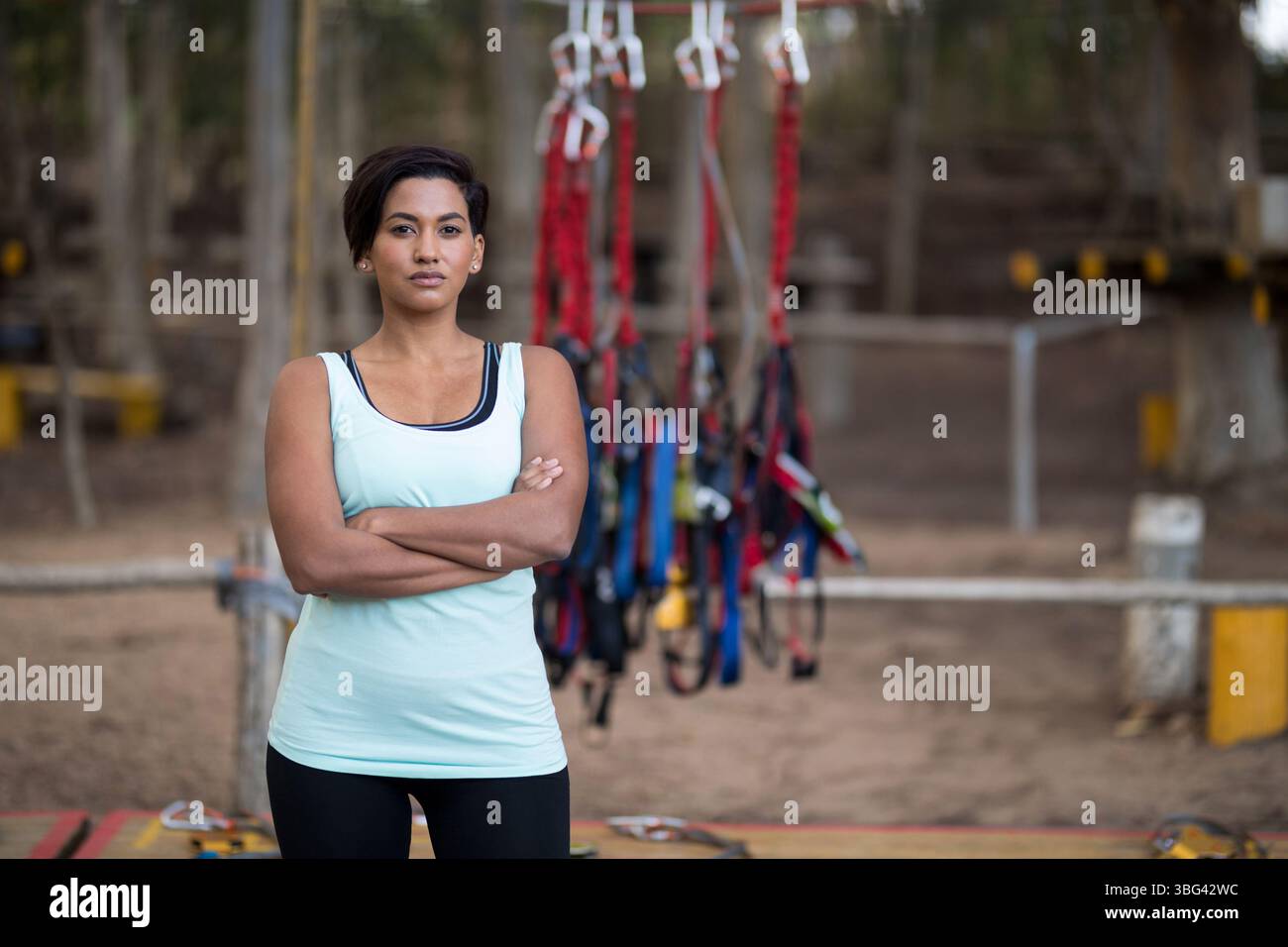 Donna in abbigliamento sportivo in piedi con le braccia incrociate di fronte all'attrezzatura di sicurezza al parco avventura aereo Foto Stock