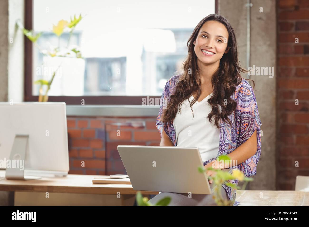 Donna in un cardigan che tiene in mano un computer portatile seduto alla scrivania con piante in ufficio, spazio per fotocopie Foto Stock