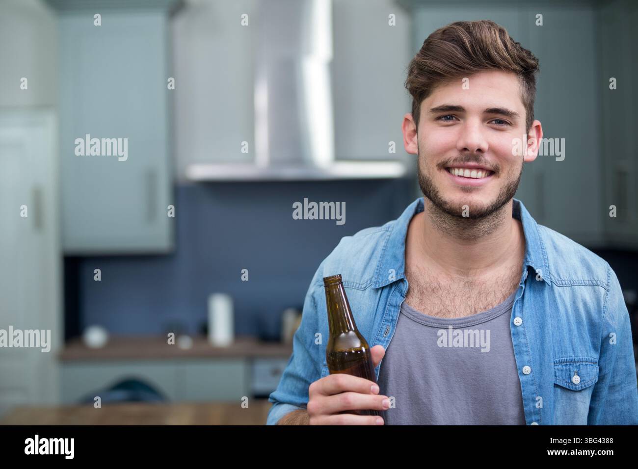 Uomo adulto che tiene in mano una bottiglia di birra e si trova nella cucina di casa con armadietti grigi, utensili, spazio fotocopie Foto Stock