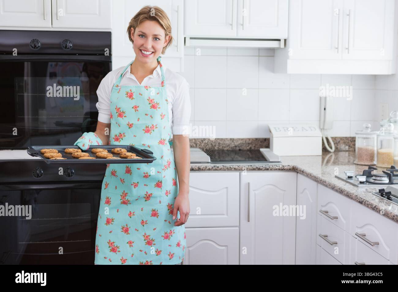 Donna che tiene in mano un vassoio per biscotti in cucina con doppio forno, con grembiule floreale pastello e guanto da forno Foto Stock