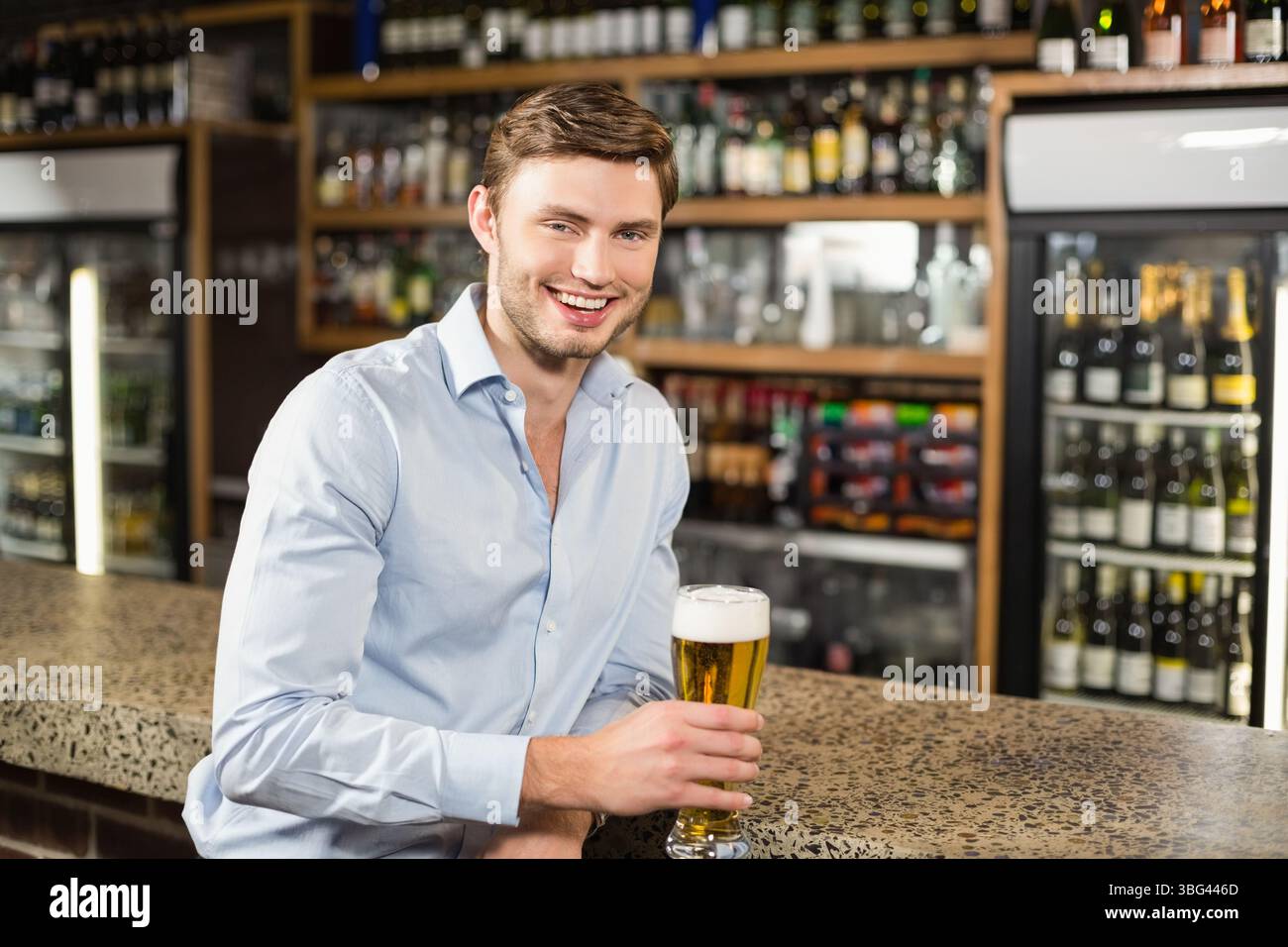 Uomo seduto al bancone del bar che tiene un bicchiere di birra, sorride vicino al refrigeratore con porta in vetro e ripiani in legno Foto Stock