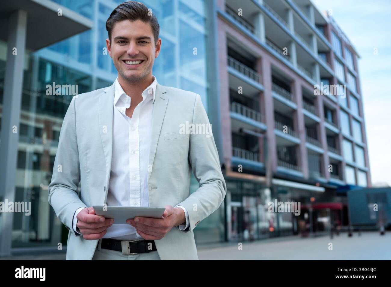 Uomo professionale che indossa un abito in piedi nella moderna piazza urbana che regge un tablet e sorride Foto Stock