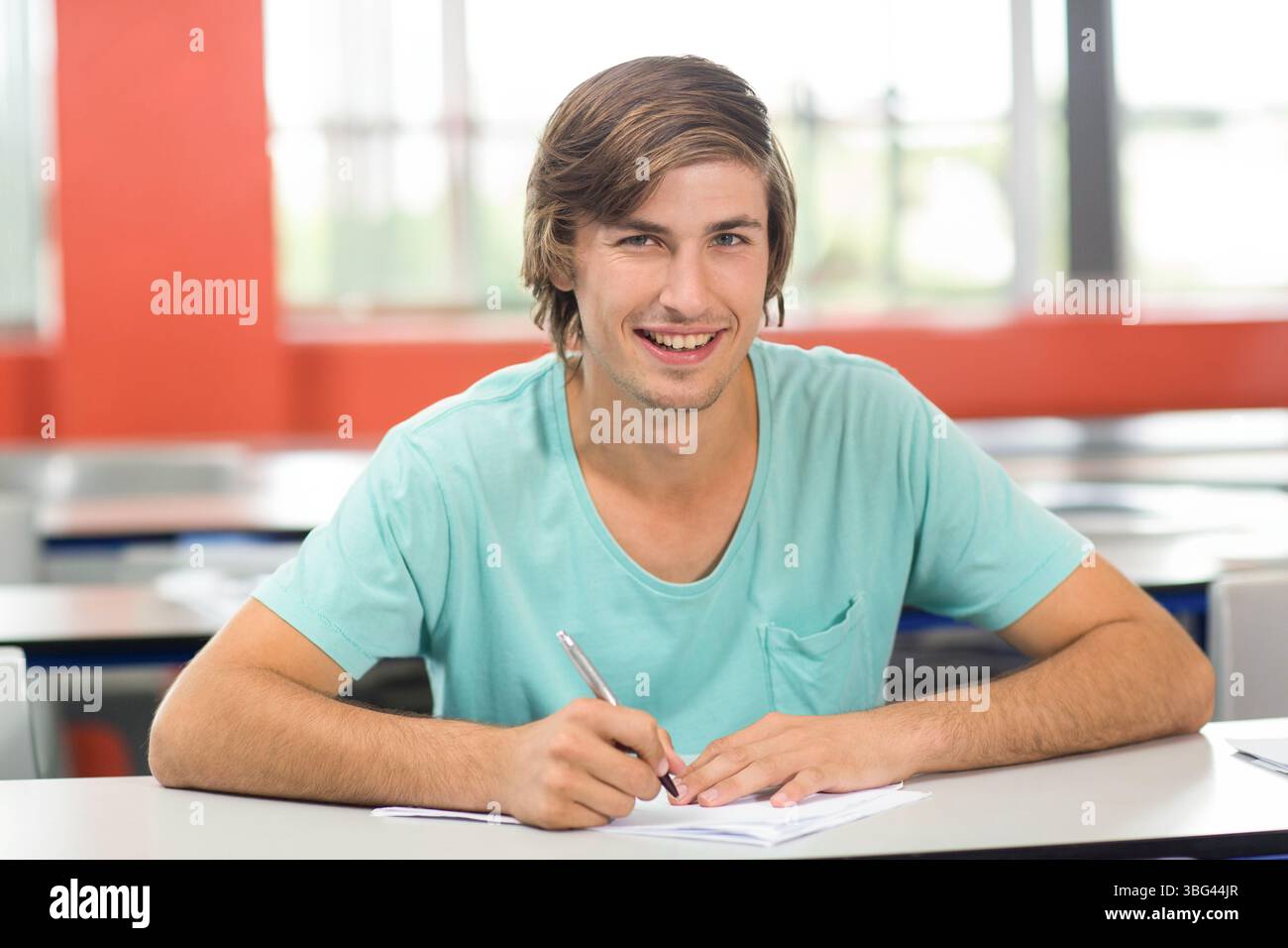 Studente di sesso maschile che tiene una penna su un notebook alla scrivania dell'aula vicino alle pareti rosse e alle grandi finestre Foto Stock