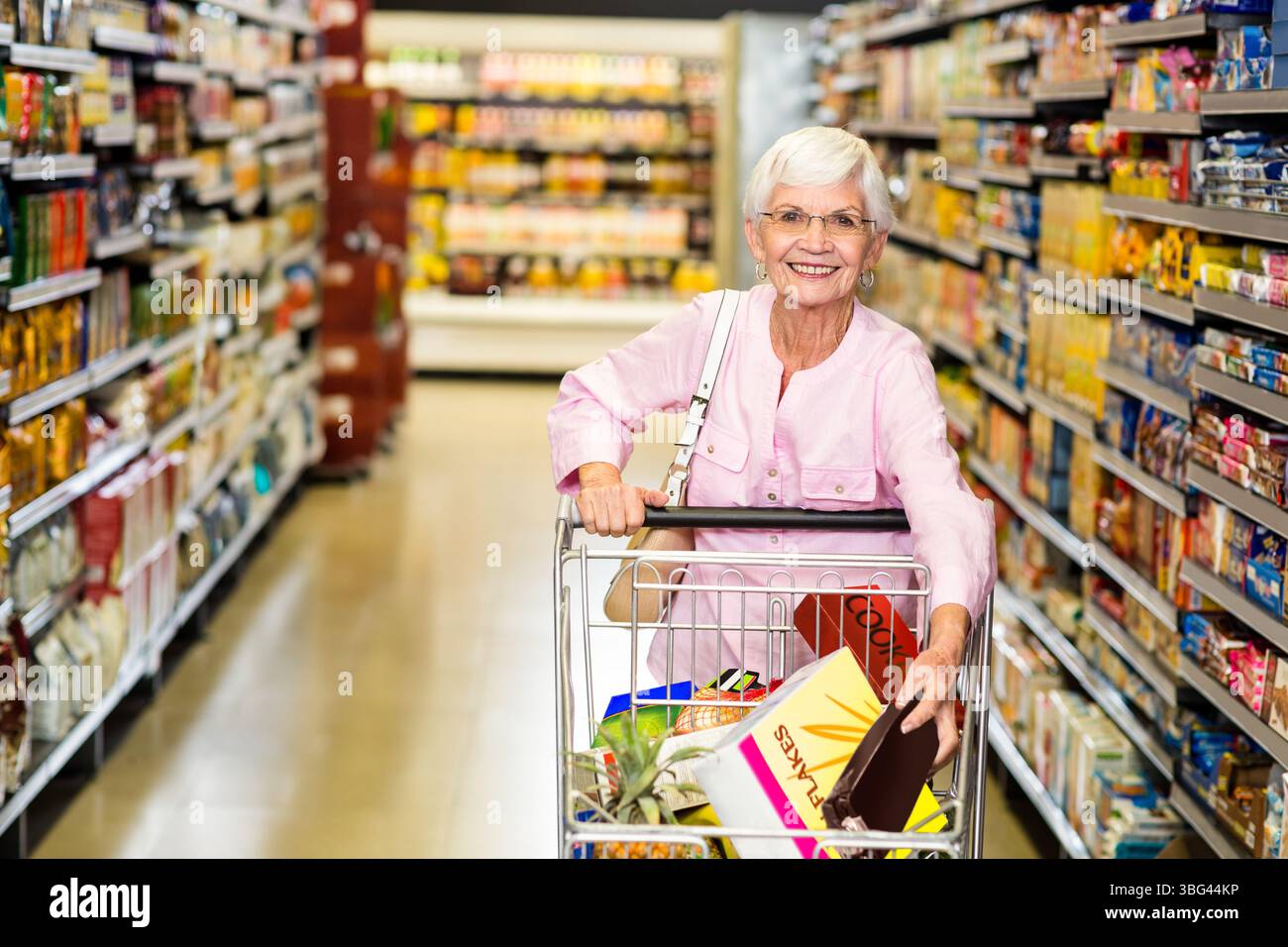 Cliente donna anziana che spinge il carrello della spesa lungo il corridoio della spesa, trasporta scatole di cereali e ananas Foto Stock