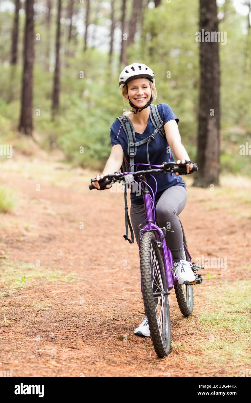Donna che cavalca una mountain bike viola lungo il sentiero della pineta con casco bianco e confezione idratante Foto Stock