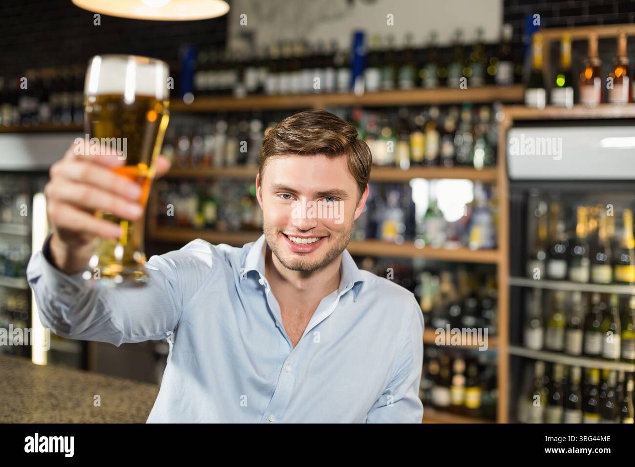 Un uomo di vent'anni che porta un bicchiere di birra pilsner con un toast al bancone del bar con scaffali per liquori Foto Stock