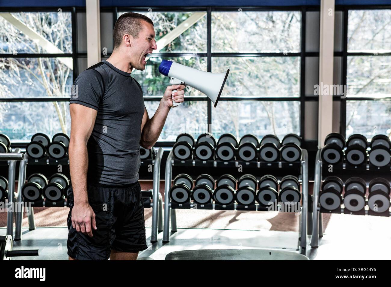 L'uomo proietta la voce attraverso il megafono bianco e blu in palestra da grandi finestre con manubri Foto Stock