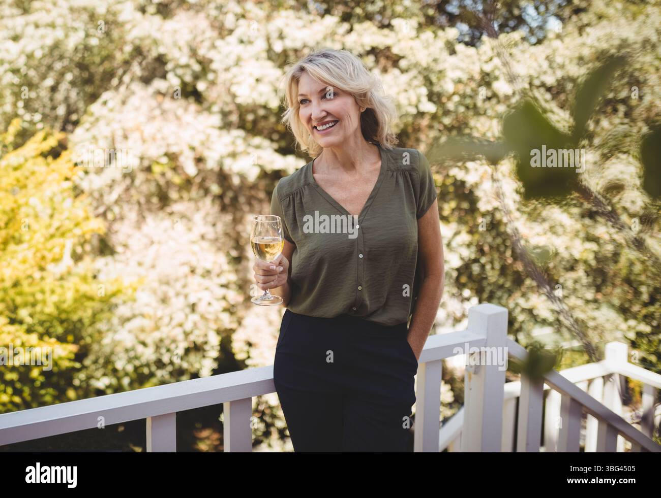 Donna in piedi sul ponte di legno nel cortile posteriore con bicchiere di vino e appoggiata alla ringhiera Foto Stock