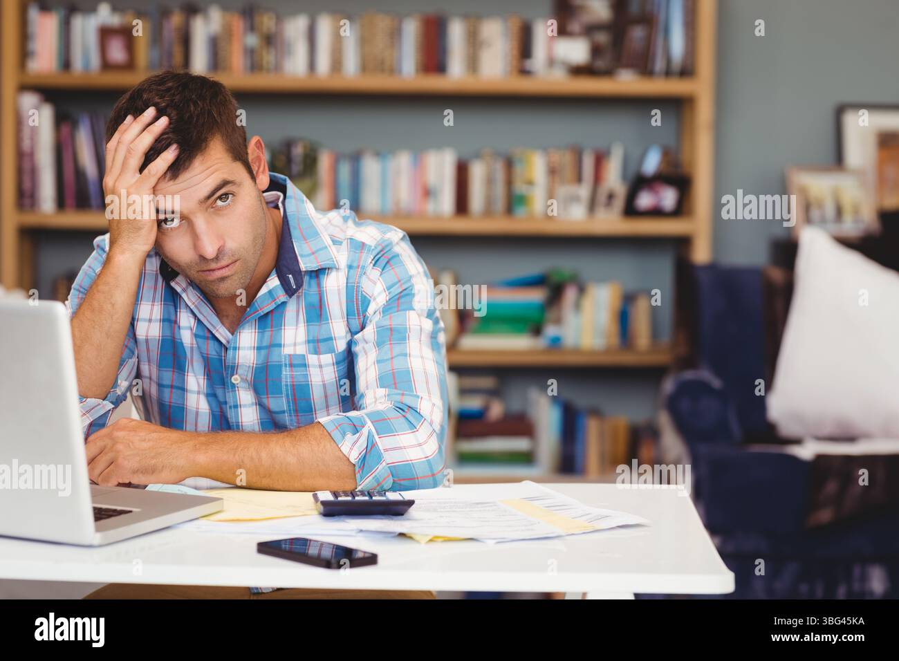 Uomo che lavora su documenti finanziari alla scrivania dell'ufficio con computer portatile, smartphone e documenti Foto Stock