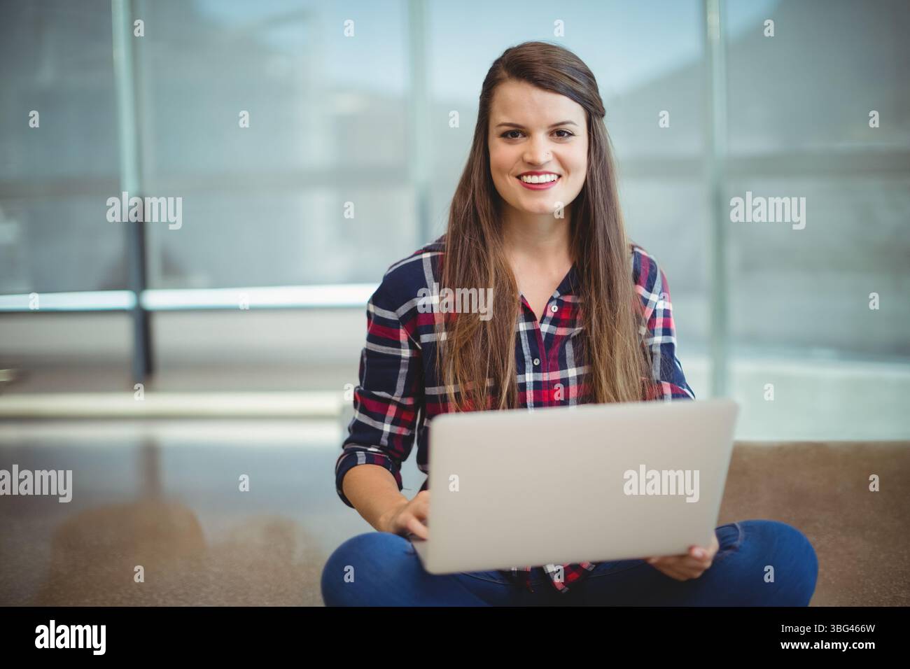 Donna che indossa camicia a quadri e jeans seduti su un pavimento lucido nella lounge dello studio utilizzando un computer portatile color argento Foto Stock