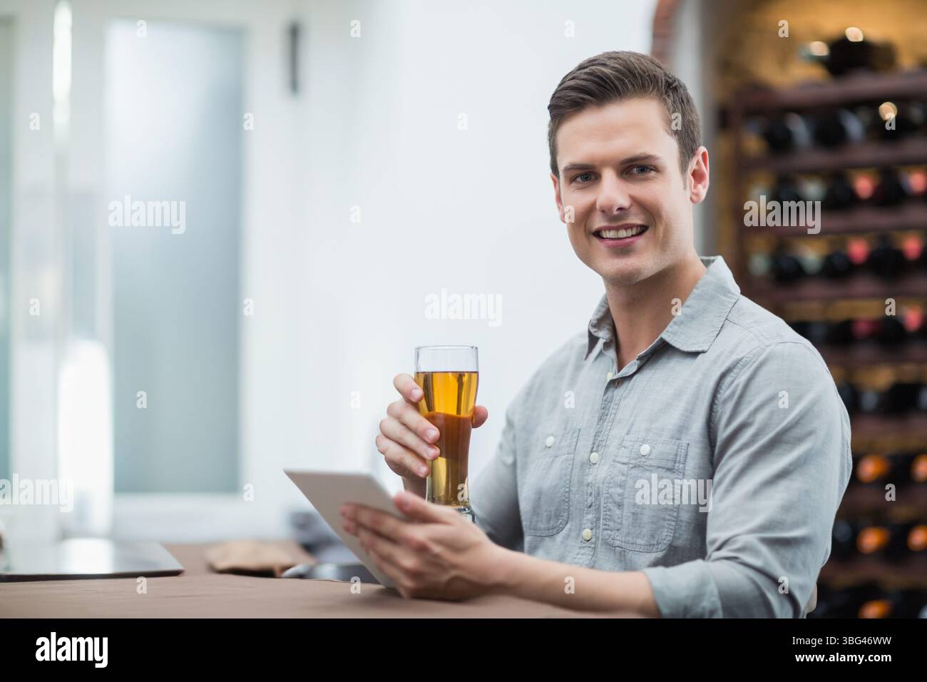 Uomo che indossa una camicia grigia seduto al bancone del bar tenendo un bicchiere di birra, utilizzando un tablet accanto alla griglia dei vini Foto Stock