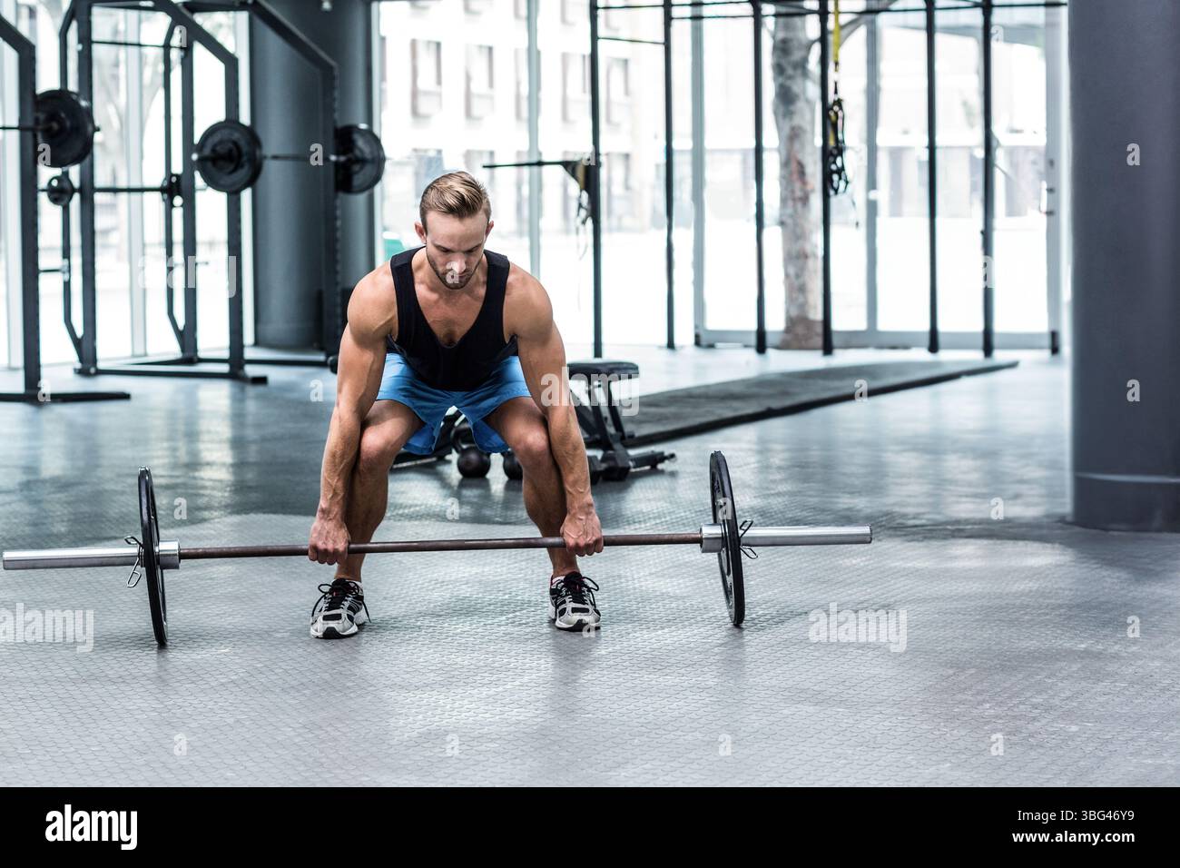 Uomo con abbigliamento sportivo che solleva una campana carica sul piano della palestra presso il centro fitness con scaffali per squat Foto Stock