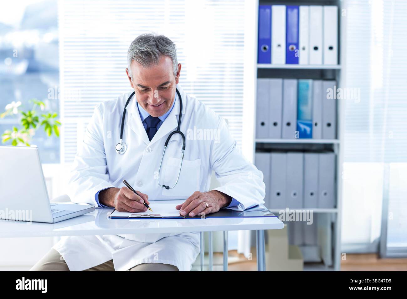 Medico di sesso maschile seduto alla scrivania scrivendo su appunti indossando stetoscopio in ufficio medico Foto Stock
